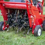 Close-up of a red mechanical core aerator machine pulling dirt plugs from a lawn to relieve soil compaction.