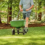 A person pushing a green broadcast spreader to apply grass seed and fertilizer across a residential lawn.