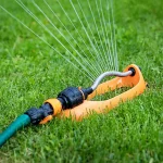 An bright orange oscillating lawn sprinkler attached to a green garden hose actively watering a yard.