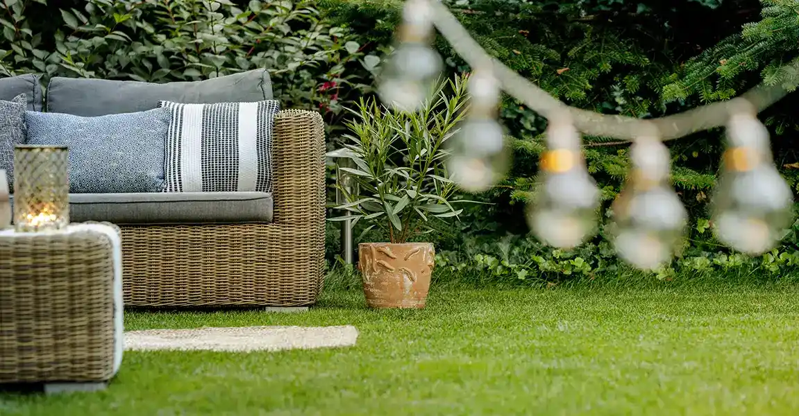 An inviting backyard outdoor living space featuring wicker patio furniture resting on a vibrant, well-maintained green lawn, framed by lush landscaping and blurred string lights.