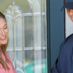 A friendly pest control professional in a dark uniform and cap smiling while speaking with a mother and her young daughter at the front door of their home.