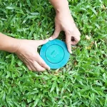 Two hands carefully placing a circular green plastic termite bait station lid into a hole in a lush green lawn.