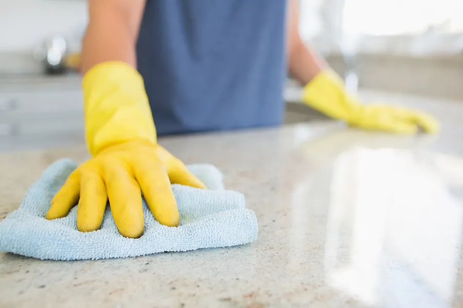 A person wearing bright yellow rubber gloves uses a light blue cloth to wipe down a polished granite countertop.