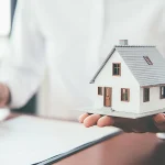 A person holding a small model of a white house in their palm while holding a pen over insurance paperwork on a clipboard, symbolizing home protection and policy coverage.