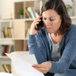 A woman looking concerned and stressed while sitting in her living room, holding a white document in one hand and talking on a smartphone with the other.