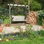 A backyard patio featuring a wooden porch swing and a large stack of firewood placed directly on the ground near the patio's edge.