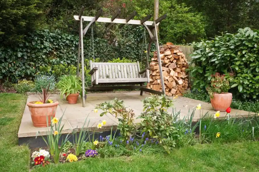 A backyard patio featuring a wooden porch swing and a large stack of firewood placed directly on the ground near the patio's edge.