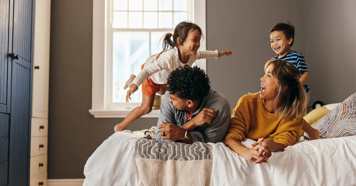 A happy family of four playing and laughing together on a bed in a bright, modern bedroom, representing a safe and protected home environment.