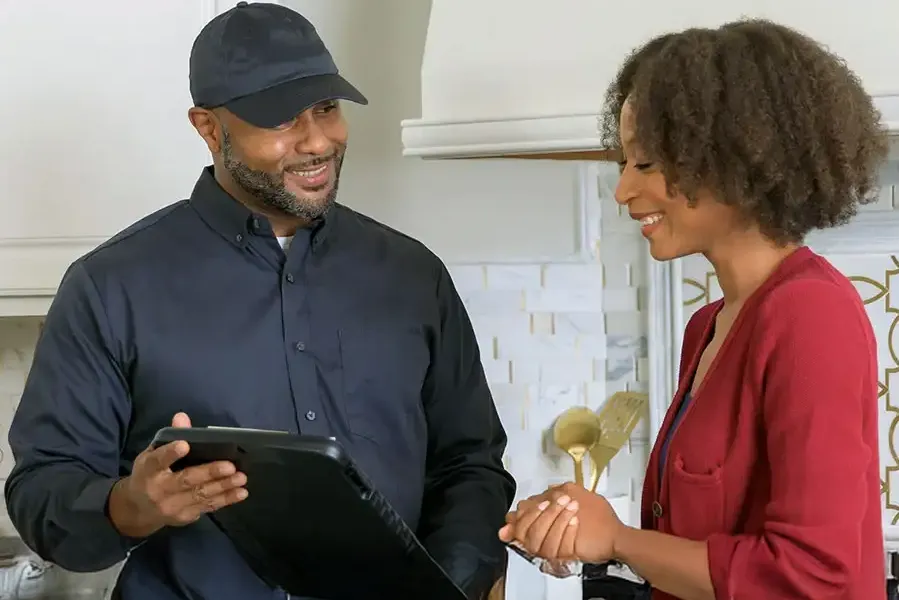 A friendly pest control technician in a black uniform and cap shows information on a tablet to a woman in a white kitchen.