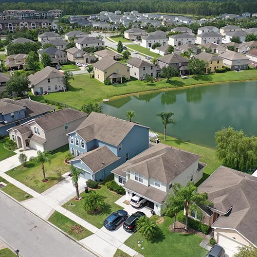 An aerial view of a large Florida subdivision centered around a man-made lake. The houses are mostly two-story single-family homes with gray or tan siding. Several cars are parked in driveways, and the neighborhood is surrounded by dense green forest in the distance.