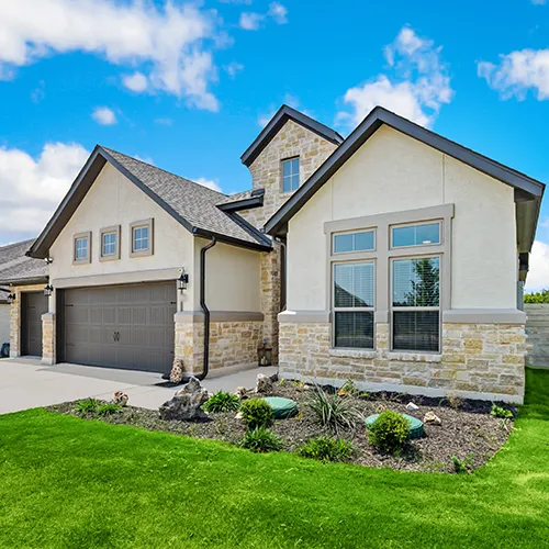 A modern, single-story home featuring a combination of cream-colored stucco and light-colored stone masonry. The house has a dark gray shingle roof with multiple gables and a large, dark brown garage door. The front yard includes a small mulched garden bed with green shrubs and decorative rocks set against a vibrant green lawn.