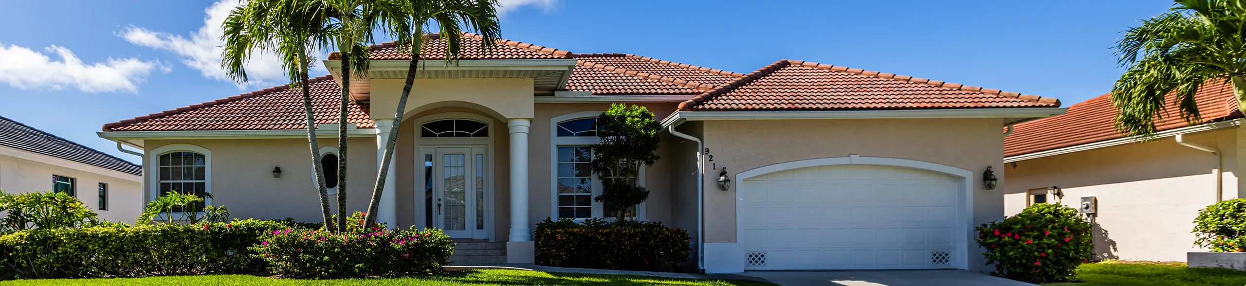A wide-angle shot of a luxury single-story villa with a light beige exterior and a red terracotta tile roof. Two tall palm trees stand prominently in the center of a lush, bright green front lawn. The house features a large white garage door and an arched entryway with white columns.