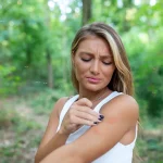 Person inspecting mosquito bites on their arm in a foresty area surrounded by mosquitoes